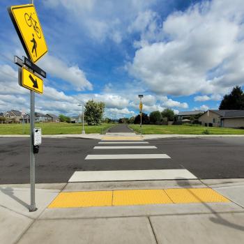 "Reedville Trail street crossing with rapid rectangular flashing beacon to the left, crosswalk looking north down the trail in center and blue sky with clouds behind the trail and greenery.""