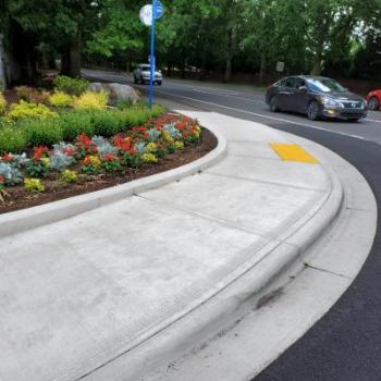 "ADA-compliant sidewalk and curb ramp at an intersection on the right of the image with landscaping to the upper left, a bus stop sign next to the landscaping and two vehicles on the street in the background."